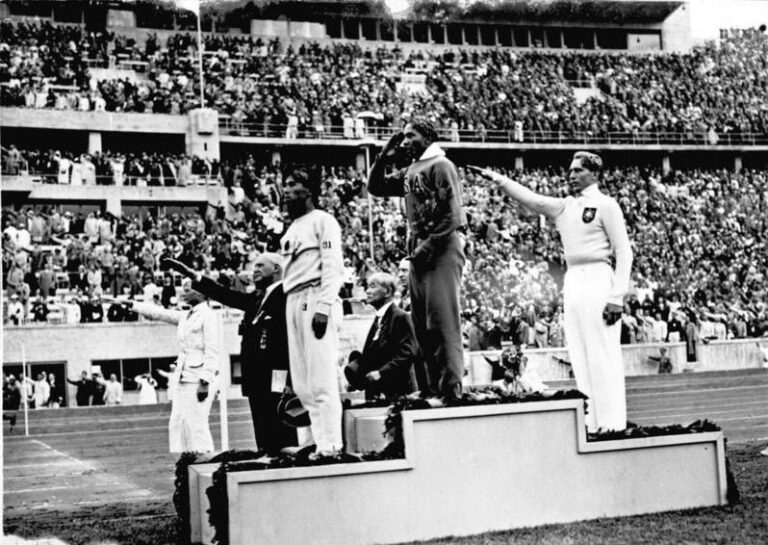 Pamětihodná fotografie po závodu ve skoku do dálky. Uprostřed na nejvyšším stupínku Jesse Owens, napravo stříbrný Long. FOTO: Bundesarchiv, Bild 183-G00630 / Unknown authorUnknown author / CC-BY-SA 3.0, CC BY-SA 3.0 DE <https://creativecommons.org/licenses/by-sa/3.0/de/deed.en>, via Wikimedia Commons