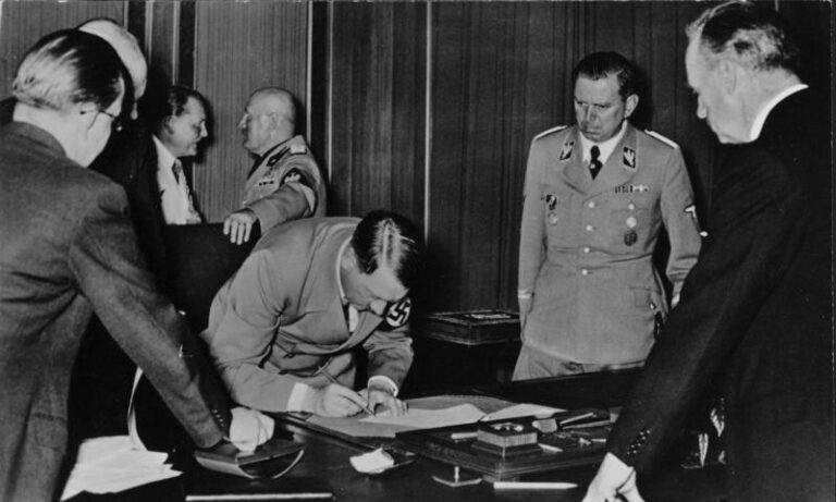 Adolf Hitler při podepisování Mnichovské dohody dne 29. září 1938. Vzadu v pozadí stojí říšský maršál Hermann Göring, který diskutuje s italským vůdcem Benitem Mussolinim. Vedle Hitlera stojí v uniformě SS jeho pobočník Julius Schaub a muž vpravo v obleku opřený o stůl je říšský ministr zahraničí Joachim von Ribbentrop.