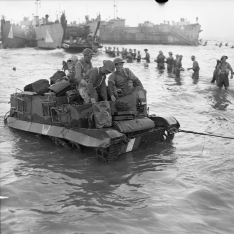 A Universal carrier is towed ashore, as troops unload ammunition from a landing craft in the background, 10 July 1943.